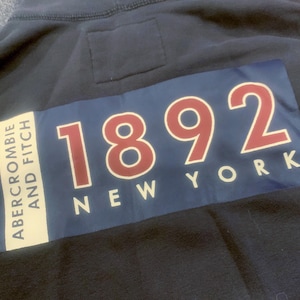 May include: Navy blue hoodie with a rectangular patch featuring the text "1892 NEW YORK" in red and white, with "Abercrombie and Fitch" printed vertically on the left side.