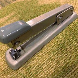 May include: A vintage gray stapler with a light blue top. The stapler has a metal body and a curved design. The number "56" is printed on the top. The stapler is on a green and tan patterned surface.