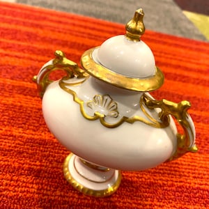 K&ouml;nnte beinhalten: Eine wei&szlig;e und goldfarbene dekorative Urne mit Deckel, Griffen und Sockel. Die Urne hat goldfarbene Verzierungen und Akzente, einschlie&szlig;lich einer goldenen Spitze auf dem Deckel. Die Urne steht auf einer orangefarbenen und roten Oberfl&auml;che.