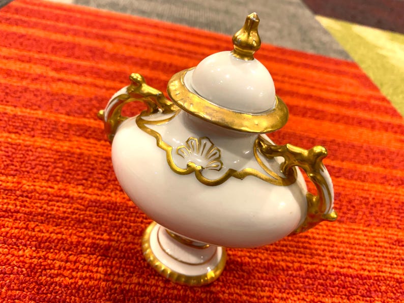 K&ouml;nnte beinhalten: Eine wei&szlig;e und goldfarbene dekorative Urne mit Deckel, Griffen und Sockel. Die Urne hat goldfarbene Verzierungen und Akzente, einschlie&szlig;lich einer goldenen Spitze auf dem Deckel. Die Urne steht auf einer orangefarbenen und roten Oberfl&auml;che.