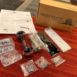May include: A Nostalgic Warehouse door hardware set including a chrome finish round knob, a rectangular backplate, a latch mechanism, mounting screws, and installation instructions in a branded cardboard box.