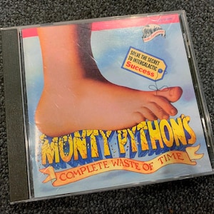 May include: A CD case featuring Monty Python's Complete Waste of Time artwork. The cover displays a large foot, a blue sky background, and a yellow tag with the text Solve the Secret to Intergalactic Success tied to a toe with string.