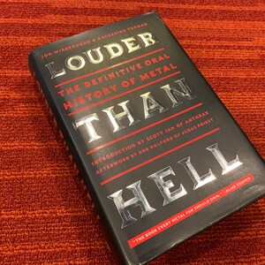 Puede incluir: Un libro de tapa dura negro titulado "Louder Than Hell: The Definitive Oral History of Metal" con letras plateadas y detalles en rojo. El libro est&aacute; sobre una superficie texturizada de color rojo anaranjado. El libro trata sobre la historia del metal.