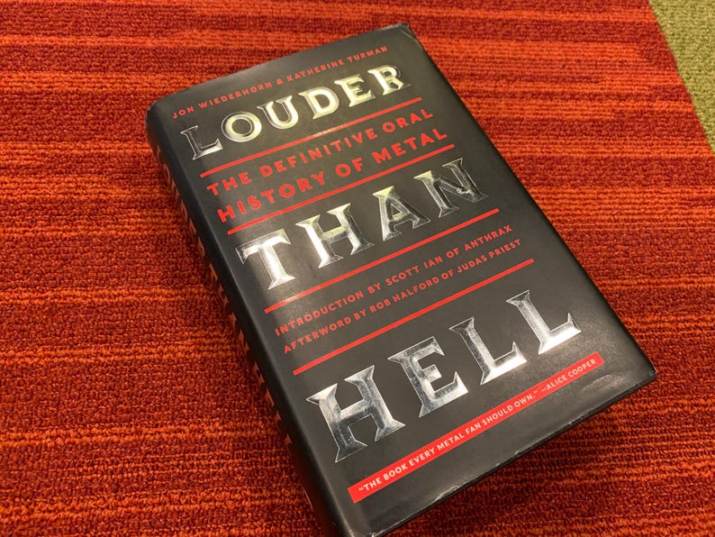 Puede incluir: Un libro de tapa dura negro titulado "Louder Than Hell: The Definitive Oral History of Metal" con letras plateadas y detalles en rojo. El libro est&aacute; sobre una superficie texturizada de color rojo anaranjado. El libro trata sobre la historia del metal.