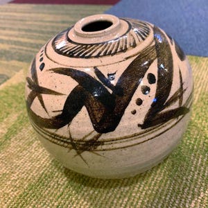 May include: A round, off-white ceramic vase with a small opening at the top. The vase is decorated with dark brown, abstract brushstrokes and patterns. The design includes a band of lines near the top and a series of dots.