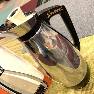 May include: A silver and black stainless steel thermal carafe with a black handle and a gold inscription that reads "Sunflower".