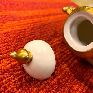 K&ouml;nnte beinhalten: Ein wei&szlig;er Keramikdeckel mit einer goldfarbenen Spitze liegt neben einem wei&szlig; und goldfarben verzierten Keramikbeh&auml;lter. Der Beh&auml;lter und der Deckel befinden sich auf einer rot-orangen strukturierten Oberfl&auml;che. Der Deckel ist eif&ouml;rmig.