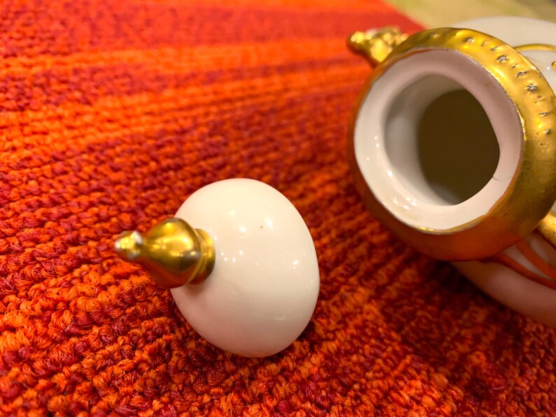 K&ouml;nnte beinhalten: Ein wei&szlig;er Keramikdeckel mit einer goldfarbenen Spitze liegt neben einem wei&szlig; und goldfarben verzierten Keramikbeh&auml;lter. Der Beh&auml;lter und der Deckel befinden sich auf einer rot-orangen strukturierten Oberfl&auml;che. Der Deckel ist eif&ouml;rmig.