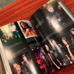 Puede incluir: Un libro abierto con fotograf&iacute;as de una banda de heavy metal. Las im&aacute;genes incluyen fotos de actuaciones en vivo y retratos de los miembros de la banda. El libro est&aacute; abierto sobre una superficie a rayas rojas y naranjas.