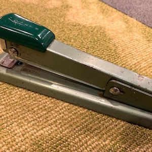 May include: A vintage green metal stapler with a green plastic top. The stapler has a silver lever and a silver base.