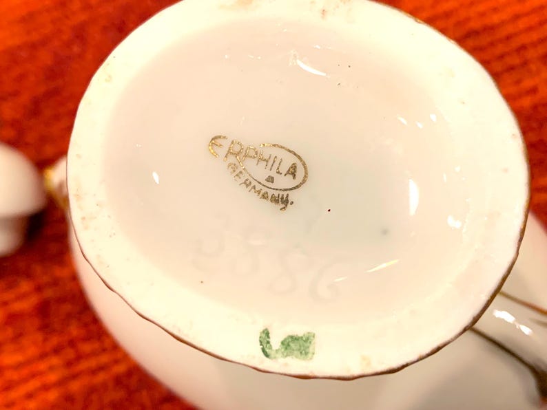 K&ouml;nnte beinhalten: Nahaufnahme des Bodens einer wei&szlig;en Porzellan-Sahnek&auml;nnchen mit Goldrand. Die Worte "ERPHILA GERMANY" sind in goldener Schrift. Die Nummer "3586" ist in das Porzellan ge&auml;tzt.