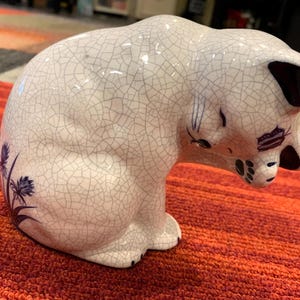 May include: A ceramic cat figurine with a white crackle glaze finish. The cat is in a seated, grooming posture with its head bowed. It features dark blue floral accents on its side, dark blue ear interiors, and painted facial details.