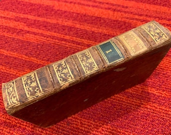 Könnte beinhalten: Ein antikes Buch mit einem abgenutzten, braunen Ledereinband. Der Buchrücken weist goldene Schrift und dekorative Blumenmuster auf. Ein dunkelgrünes Feld mit der Zahl "I" ist sichtbar. Das Buch liegt auf einer roten, strukturierten Oberfläche.