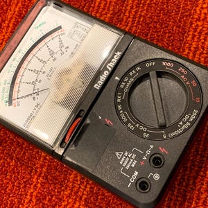 May include: A black Radio Shack multimeter with a clear plastic cover. The device has a dial with settings for ohms, ACV, and DCA. The display shows various measurements and scales. The device is on an orange textured surface.
