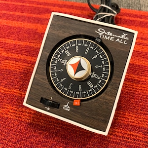 May include: An Intermatic Time All electrical timer with a wood grain finish faceplate. The device features a circular black dial with white hour markings, a central orange and black graphic, and a small orange switch labeled ON.
