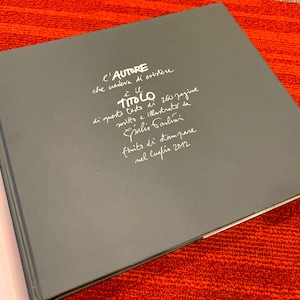 May include: A dark gray hardcover book with white handwritten-style text on the front cover. The text reads: C'AUTORE che credeva di esistere è il TITOLO di questo testo di 260 pagine scritto e illustrato da Giulio Carlini finito di stampare nel luglio 2012.