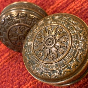 May include: Two round, metallic bronze-toned door knobs featuring an intricate, embossed pattern. The design includes a central floral motif surrounded by concentric circles of textured geometric shapes and small flower accents.