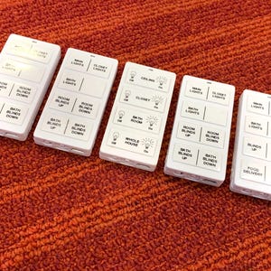 May include: Five rectangular white remote control units with black text labels for home automation. Each unit features multiple buttons for controlling lights, room blinds, and specific household functions like food delivery and bed time settings.