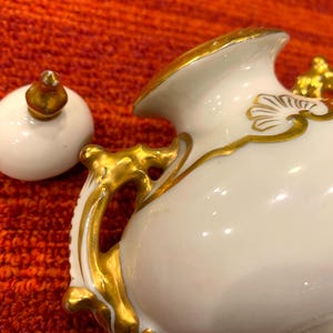 K&ouml;nnte beinhalten: Eine wei&szlig;e Porzellan-Sahnek&auml;nnchen mit goldenen Akzenten. Das Sahnek&auml;nnchen hat einen geschwungenen Griff und ein dekoratives Golddesign. Der Deckel ist wei&szlig; mit einem goldenen Knauf. Das Sahnek&auml;nnchen steht auf einer orangefarbenen Oberfl&auml;che.