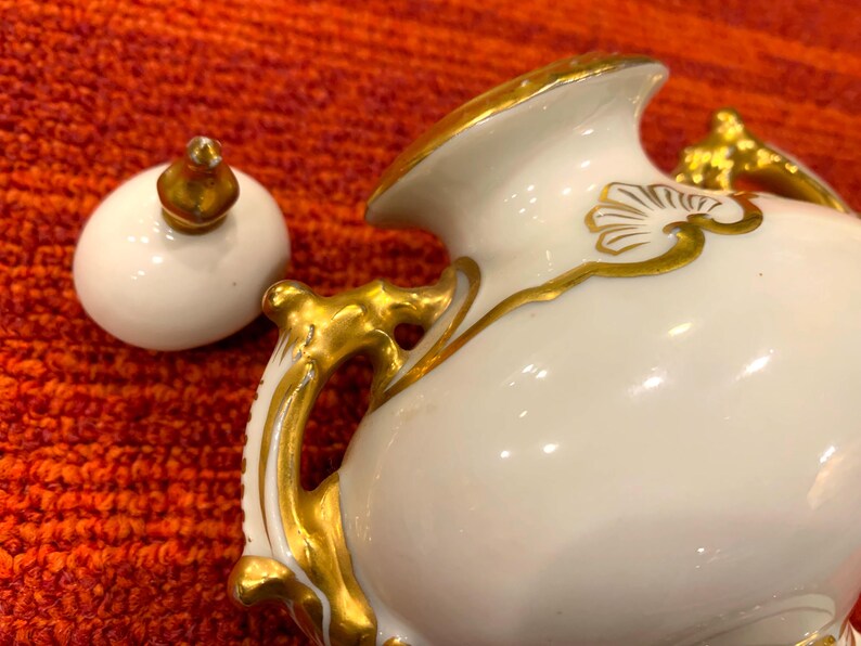 K&ouml;nnte beinhalten: Eine wei&szlig;e Porzellan-Sahnek&auml;nnchen mit goldenen Akzenten. Das Sahnek&auml;nnchen hat einen geschwungenen Griff und ein dekoratives Golddesign. Der Deckel ist wei&szlig; mit einem goldenen Knauf. Das Sahnek&auml;nnchen steht auf einer orangefarbenen Oberfl&auml;che.