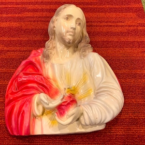 May include: A religious bust sculpture depicting a figure with long hair and a beard, wearing a white robe with a red drape. The figure holds a heart with a yellow glow. The sculpture is on a red carpet.