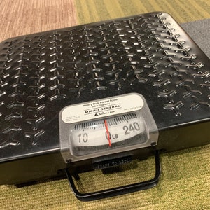 May include: Black heavy-duty floor scale with a black metal platform and a silver dial. The dial reads "MICRO GENERAL" and has markings from 10 to 240 pounds. The scale has a black handle.