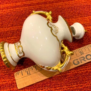 K&ouml;nnte beinhalten: Eine wei&szlig;e Porzellanvase mit goldenen Akzenten, einschlie&szlig;lich Griffen und dekorativen Mustern. Die Vase hat einen bauchigen K&ouml;rper und einen kleinen Deckel. Ein Holzlineal liegt neben der Vase.