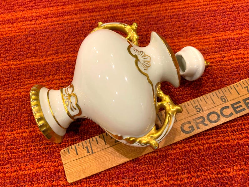 K&ouml;nnte beinhalten: Eine wei&szlig;e Porzellanvase mit goldenen Akzenten, einschlie&szlig;lich Griffen und dekorativen Mustern. Die Vase hat einen bauchigen K&ouml;rper und einen kleinen Deckel. Ein Holzlineal liegt neben der Vase.