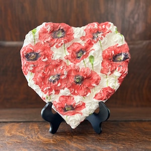 May include: A white heart-shaped ceramic plaque with a raised design of red poppies. The plaque is on a black stand.