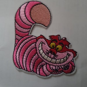 May include: Pink and red embroidered iron-on patch of the Cheshire Cat from Alice in Wonderland. The cat is grinning with a wide, toothy smile.