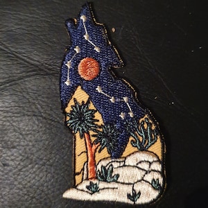 Pode incluir: Etiqueta bordada com um lobo azul a uivar para a lua vermelha com uma constelação de estrelas. O lobo está silhuetando-se contra uma paisagem desértica com palmeiras e cactos.