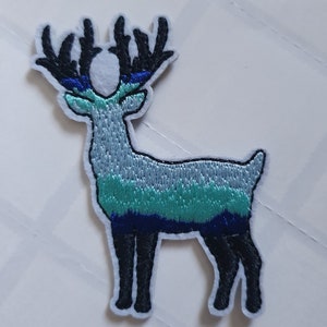 Puede incluir: Un parche bordado de hierro con un ciervo con cuernos, en tonos azules y verdes. El ciervo está de pie sobre un fondo blanco.
