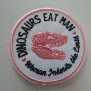 May include: Pink and white embroidered patch with the text "Dinosaurs Eat Man, Woman Inherits the Earth" and a pink dinosaur skull graphic.