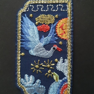 May include: Embroidered patch featuring a blue bird in flight against a dark blue background. The bird is surrounded by gold stars, clouds, and a red and orange sun. The patch is edged in gold thread.