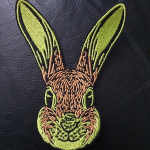 Puede incluir: Parche bordado verde y marrón de la cara de un conejo. El conejo tiene orejas grandes y una nariz marrón y esponjosa.