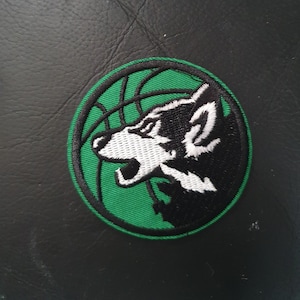 Könnte beinhalten: Grün-schwarzer gestickter Aufnäher mit einem Wolfskopf in einem Basketball. Der Wolfskopf ist weiß mit schwarzen Akzenten.