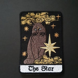 Puede incluir: Parche bordado para planchar con un gato negro con una luna creciente en la frente, sentado frente a una estrella dorada y nubes. El texto "The Star" está escrito debajo de la imagen.