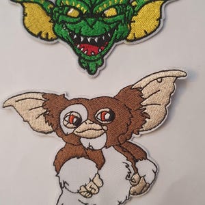 May include: Two embroidered iron-on patches featuring characters from the film Gremlins. The first patch is a green Gizmo with yellow ears and a red mouth. The second patch is a brown Mogwai with white fur and red eyes.