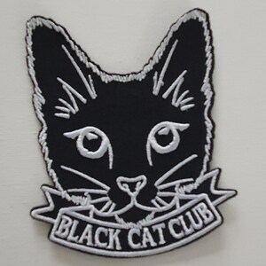 Può includere: Toppa ricamata in bianco e nero con il viso di un gatto e il testo "Black Cat Club" su uno stendardo bianco.