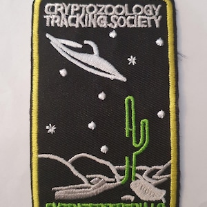 May include: A black embroidered patch with a yellow border. The patch features a white UFO flying over a green cactus and a desert landscape. The text "CRYPTOZOOLOGY TRACKING SOCIETY" is at the top and "EXTRATERRESTRIALS" is at the bottom.