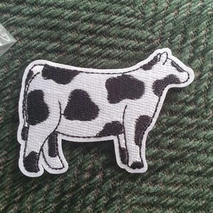 May include: A white and black embroidered patch of a cow with black spots. The cow is standing on all four legs and is facing to the right.