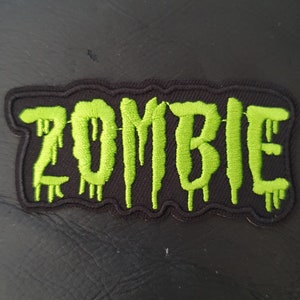 Puede incluir: Parche de tela negro con la palabra "ZOMBIE" bordada en verde en letras que gotean.