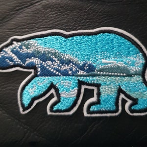 May include: An embroidered patch depicting a polar bear standing on a snowy landscape. The bear is outlined in white and features blue and white embroidery.