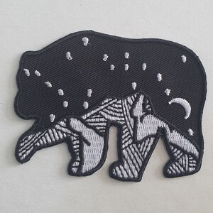 以下が含まれることがあります： 星が散りばめられた夜空を背景にした、黒と白の刺繍パッチ。クマは白で縁取られ、背中に三日月があります。