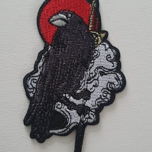 Könnte beinhalten: Schwarzer gestickter Patch mit einem Raben, der auf einem Schwert sitzt, mit einer roten Sonne dahinter. Der Rabe blickt nach links und das Schwert zeigt nach oben. Der Hintergrund ist weiß mit einem weißen Wolkenmuster.