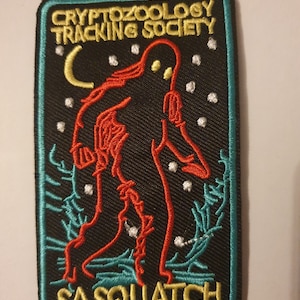 Puede incluir: Un parche bordado negro con una figura de sasquatch roja de pie en un bosque con una luna creciente y estrellas. El texto "Cryptozoology Tracking Society" está por encima del sasquatch y "Sasquatch" está debajo de la figura. El texto "Maidenvoyageclothingco." está en la parte inferior del parche.