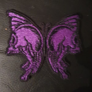 Könnte beinhalten: Lilafarbener gestickter Schmetterlings-Patch mit Totenkopfdetails. Die Schmetterlingsflügel sind in Form von Totenköpfen gestaltet.