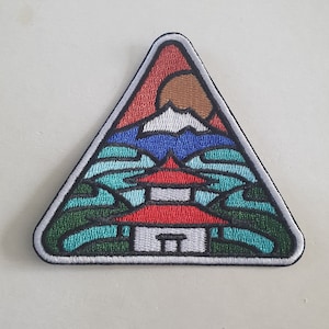 May include: A triangular embroidered patch featuring a red, white and blue mountain scene. The mountain is surrounded by green and blue waves. A small white building with a red roof sits at the base of the mountain.