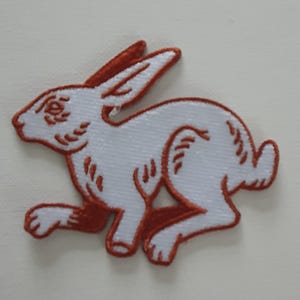 Puede incluir: Parche de conejo blanco bordado con contorno marrón, corriendo hacia la derecha. El parche está hecho de fieltro y tiene un borde cosido.