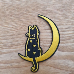 Puede incluir: Gato negro bordado sentado en una luna creciente amarilla. El gato tiene un patrón estrellado en su cuerpo.